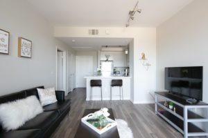 Interior view of living and TV area in student apartment, as well as kitchen with stainless steel appliances and high countertop with barstools.