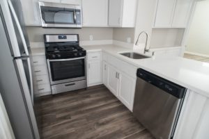 Apartment U shaped kitchen with white peninsula counter facing the living area, white cabinets and drawers, wood plank flooring, and stainless-steel appliances including: dishwasher, undermounted sink, oven and stove combo with microwave above, and single door refrigerator with top freezer